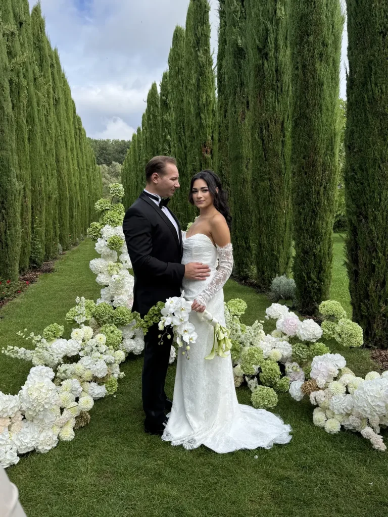 Brautpaar in eleganter Hochzeitsmode steht zwischen hohen Zypressen in einer luxuriösen Gartenkulisse mit weißen Blumenarrangements und romantischer Atmosphäre.